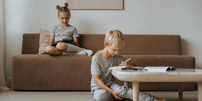 A boy studies while a girl relaxes on the sofa, both in a comfortable indoor room setting.